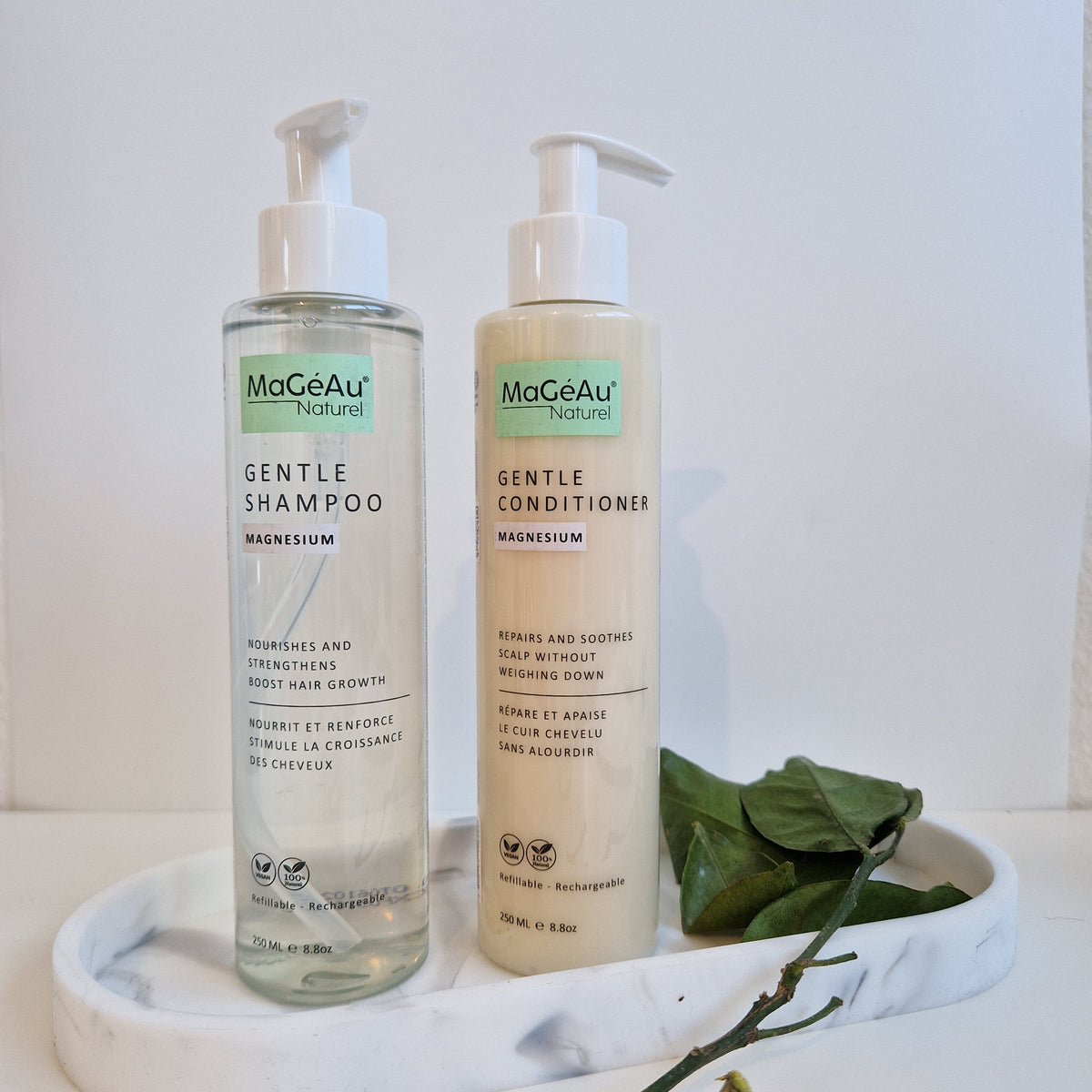 Two bottles of Mageau naturel magnesium Gentle Shampoo and Gentle Conditioner on a white surface with green leaves.